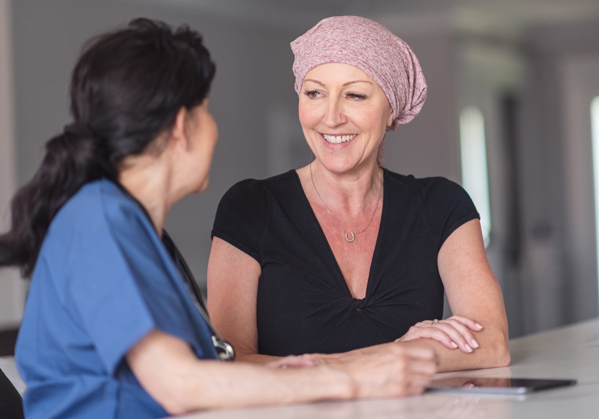 An caucasian woman with cancer is meeting with her female. They are seated next to each other in a clinic. The doctor is showing the patient test results on an electronic wireless tablet. The patient is asking the doctor questions about her treatment and recent test results. She is smiling because her doctor has given her good news.